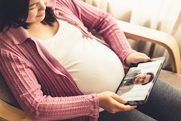 Close-up image of a pregnant woman sitting on a sofa with tablet on her lap. Health care provider appears on screen of tablet. 